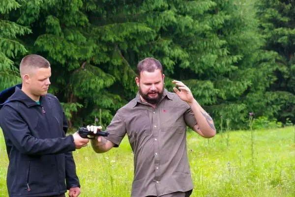 Zwei Personen stehen in einer Wiese, eine übergibt eine Pistole an die andere