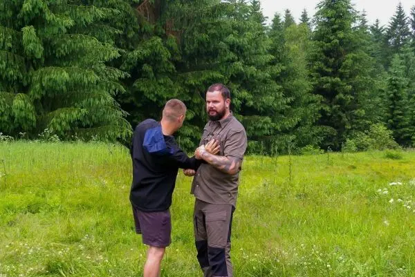 Zwei Personen üben eine körperliche Technik in einer grünen Wiese mit Bäumen im Hintergrund