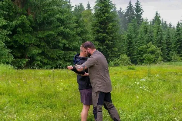 Zwei Personen üben eine Technik im hohen Gras, umgeben von Bäumen