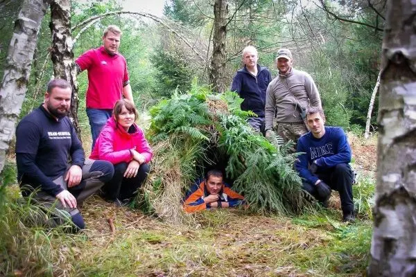 Gruppe steht vor einer selbstgebauten Tarp-Notunterkunft aus Ästen und Farnen