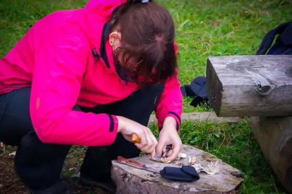 Holzstück wird mit einem Messer bearbeitet, Werkzeuge liegen auf einem Baumstamm