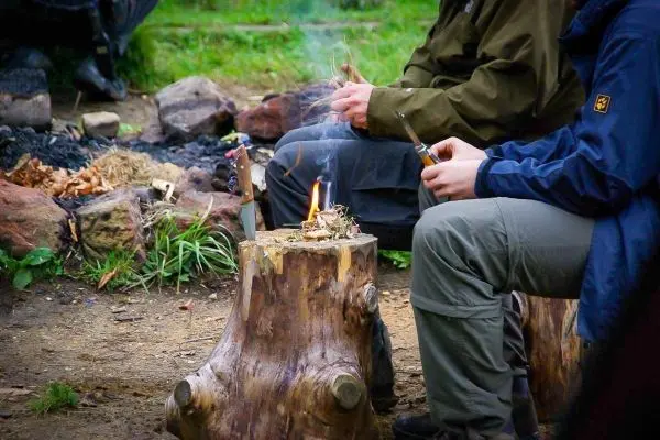 Kleine Flamme brennt auf einem Holzstamm, während zwei Personen mit Messern arbeiten