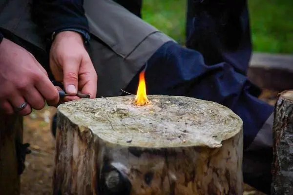 Kleine Flamme glimmt auf einem Holzscheit, während eine Hand Zunder bearbeitet