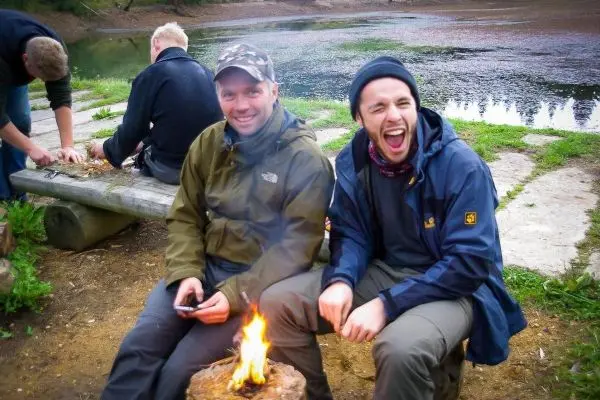 Kleines Bodenfeuer brennt auf einem Holzstamm, zwei Männer sitzen daneben