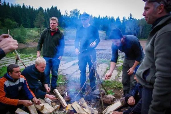Kleines Bodenfeuer mit Rauch und Holzscheiten, mehrere Personen um das Feuer versammelt