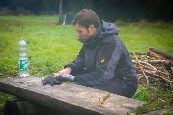 Mann sitzt an einem Holzstamm und bearbeitet mit einem Messer einen Stock