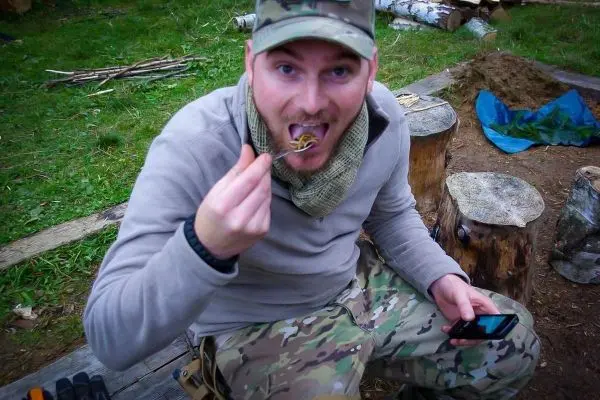 Mann sitzt auf Holzscheit und isst mit Löffel aus einer Schüssel