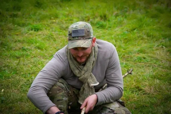 Mann sitzt im Gras und bearbeitet einen Stock mit einem Messer