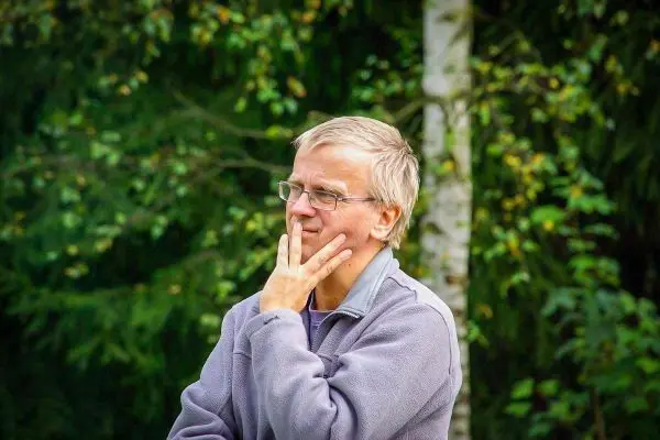 Nachdenkliche Person mit Brille und grauem Pullover vor grüner Hintergrundvegetation