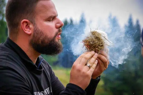 Person bläst auf ein Nest aus Gras und Zunder