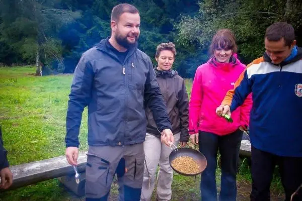 Person hält Pfanne mit Zubereitung über Feuerstelle, umgeben von weiteren Personen