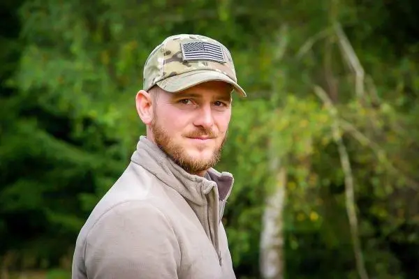 Person mit Camouflage-Mütze und grauem Pullover steht vor grüner Natur