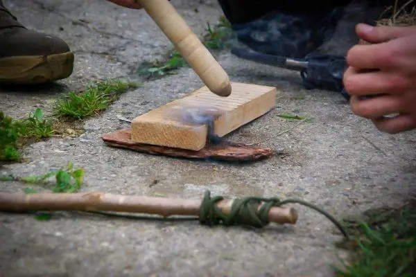 Person reibt einen Stock auf einem Holzstück beim Feuer machen