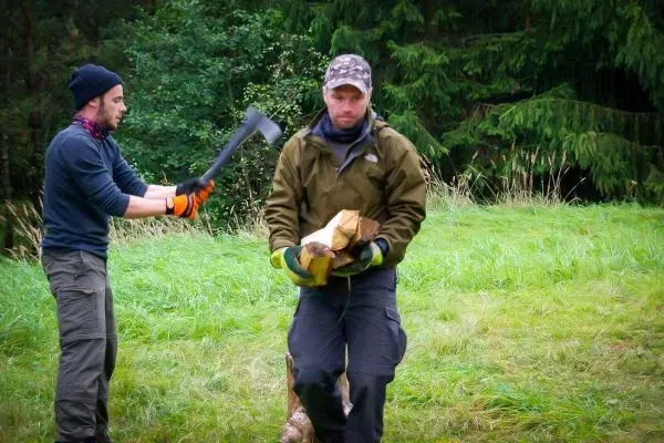 Person trägt Holz, während andere Person mit Axt arbeitet
