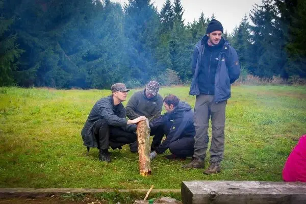 Personen bearbeiten einen Holzstamm auf einer Wiese beim Survival Training
