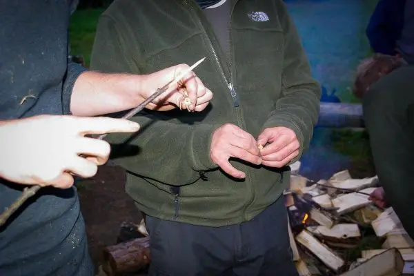 Zwei Hände halten einen Stock und ein Stück Zunder über einem Holzstapel