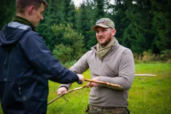 Zwei Personen halten einen Stock in einer Wiese zwischen Bäumen