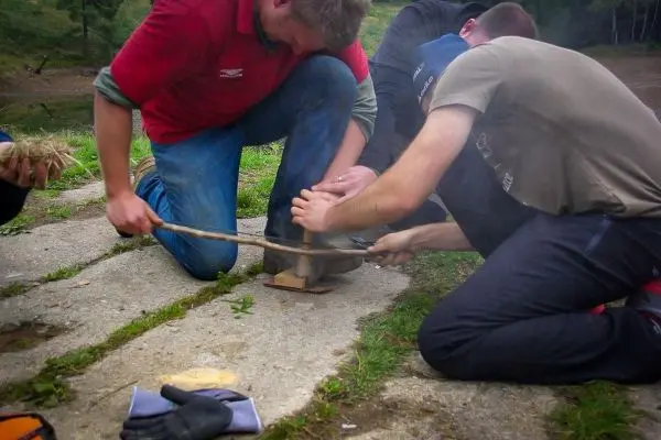 Zwei Personen nutzen einen Feuerbohrer zur Zundererzeugung auf dem Boden