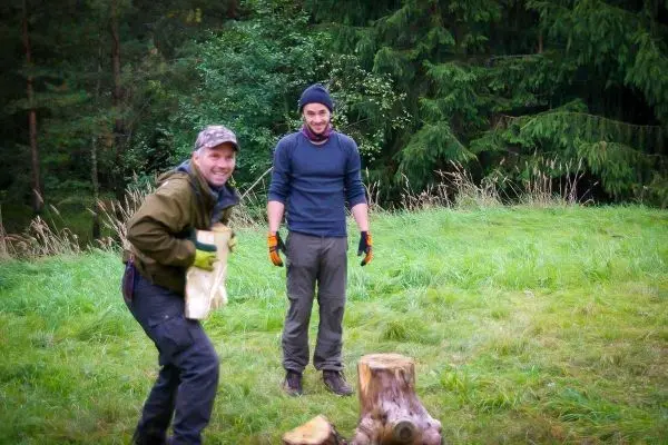 Zwei Personen stehen auf einer Wiese neben einem Baumstumpf, einer hält Holz