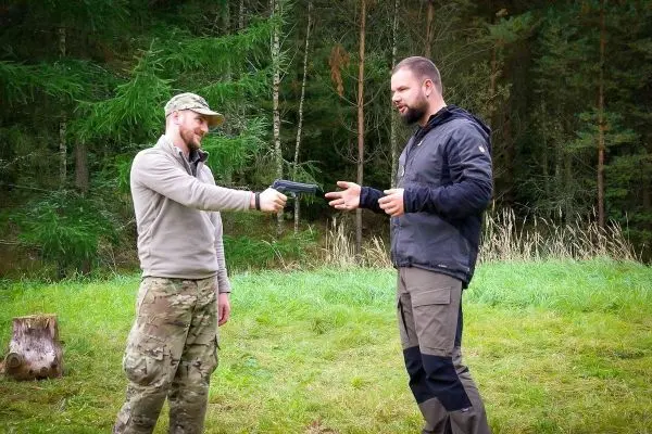 Zwei Personen stehen in einer Waldumgebung, eine Person hält eine Pistole
