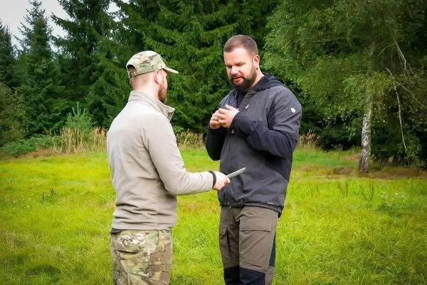 Zwei Personen stehen in einer Wiese und diskutieren, einer hält ein Messer in der Hand