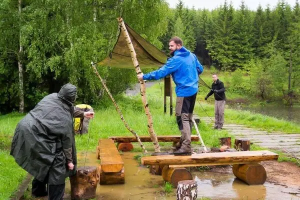 Drei Personen bauen eine Tarp-Notunterkunft mit Holzgestellen und Seilen