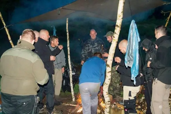 Gruppe um ein kleines Bodenfeuer versammelt, Tarp-Notunterkunft im Hintergrund