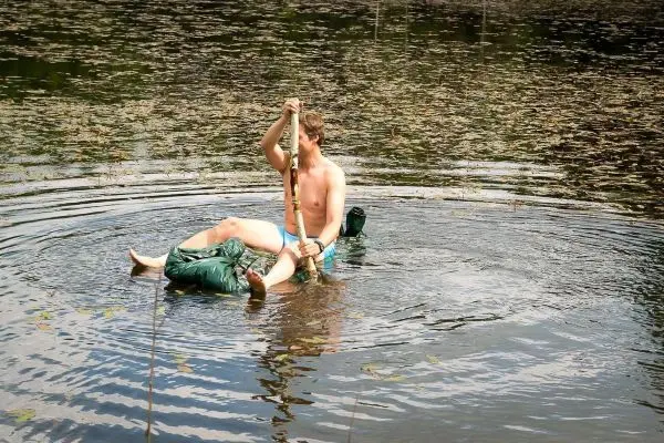 Junge sitzt auf einer improvisierten Schwimmkonstruktion im Wasser und paddelt mit einem Stock