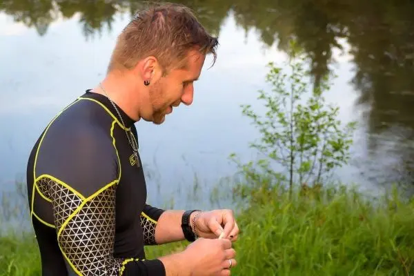Mann in Neoprenanzug hält Ausrüstung am Ufer eines Gewässers