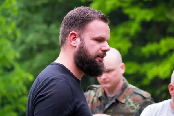 Mann mit Bart spricht in einer grünen Umgebung, andere Personen im Hintergrund