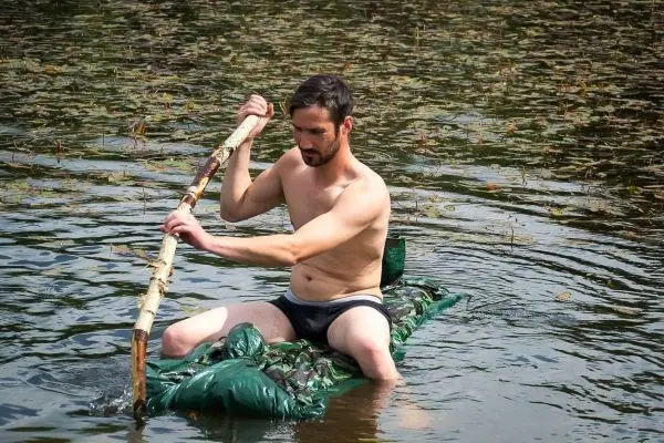Mann paddelt mit einem Stock auf einer improvisierten schwimmenden Plattform im Wasser