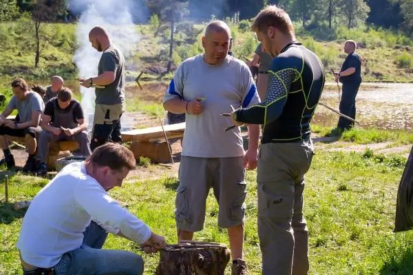 Mann schnitzt mit einem Messer an einem Holzstück, während andere am Feuer stehen