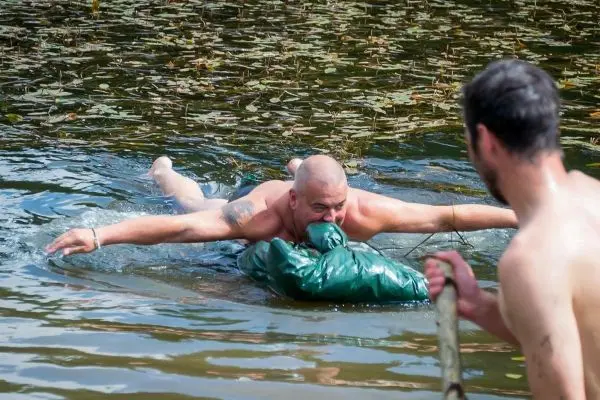 Mann schwimmt mit einem grünen Sack im Wasser, während ein anderer ihn mit einem Stock unterstützt