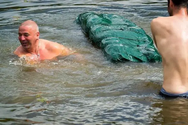 Mann schwimmt mit einer grünen Plane im Wasser, während ein anderer ihn unterstützt