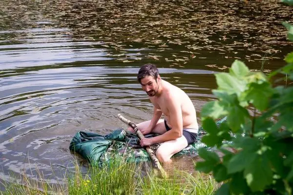 Mann sitzt auf improvisiertem Floß aus grüner Plane und Holzstöcken im Wasser