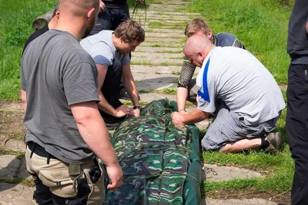 Mehrere Personen arbeiten gemeinsam an einer improvisierten Trage aus einer camouflagefarbenen Plane