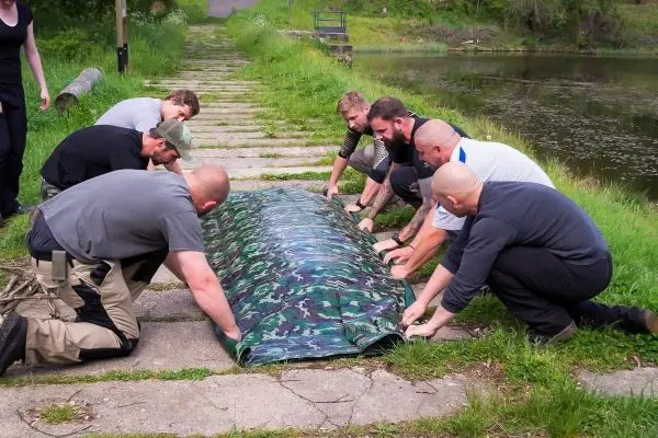 Mehrere Personen bauen gemeinsam eine Tarp-Notunterkunft am Ufer eines Gewässers auf