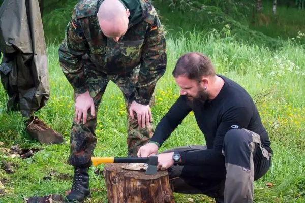 Person schnitzt mit einer Axt auf einem Baumstamm, Person schaut zu