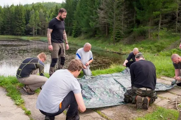 Sechs Personen bauen gemeinsam eine Tarp-Notunterkunft am Ufer eines Gewässers auf