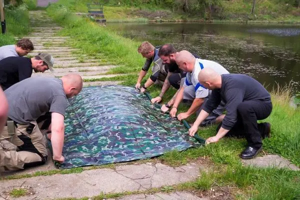 Sechs Personen spannen eine Tarp-Notunterkunft am Ufer eines Gewässers