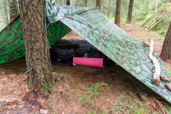 Tarp-Notunterkunft zwischen Bäumen mit Schlafunterlage und Ausrüstung darunter