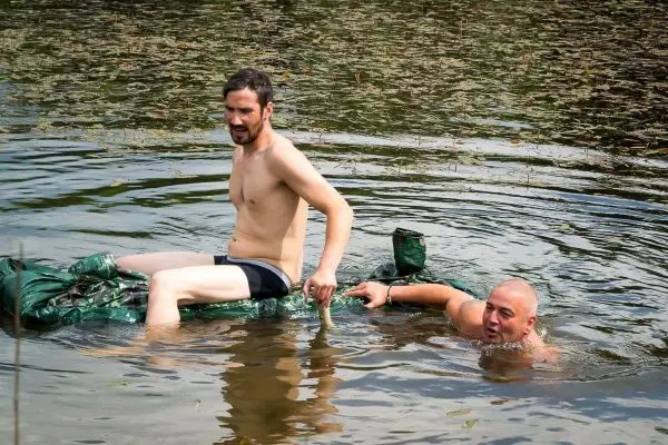 Zwei Personen auf improvisierter Schwimmhilfe im Wasser, einer hält einen Fisch
