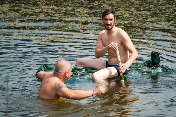 Zwei Personen im Wasser, einer sitzt auf einer grünen Unterlage und hält einen Stock