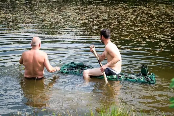 Zwei Personen sitzen auf einer improvisierten Schwimmhilfe im Wasser und halten Stöcke