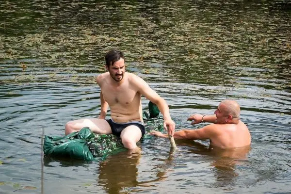 Zwei Personen sitzen in einem Gewässer, einer hält einen Stock in der Hand