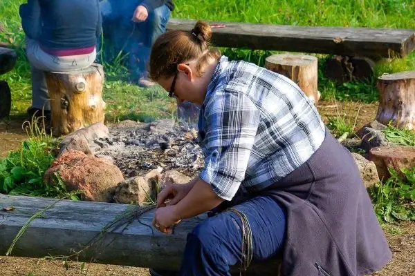 Frau bindet Pflanzenmaterial auf einem Holzstamm neben einem kleinen Feuer
