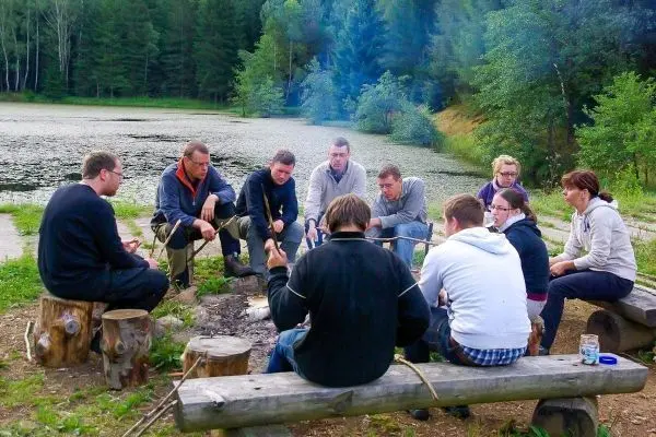 Gruppe sitzt um ein Lagerfeuer, Holzscheite als Sitzgelegenheiten, einige halten Stöcke