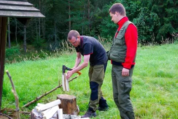 Person hackt Holz auf einem Baumstumpf, während eine andere Person zuschaut