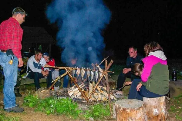 Personen sitzen um ein Feuer und grillen Fischspieße beim Überlebenstraining