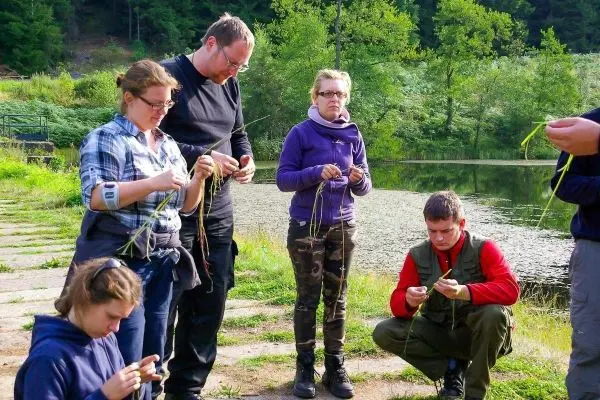 Teilnehmende flechten Pflanzenmaterialien am Ufer eines Gewässers